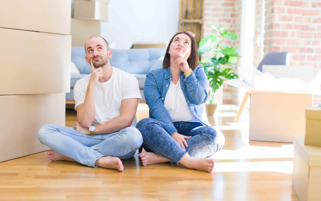 Young couple sitting on the floor arround cardboard boxes moving to a new house with hand on chin thinking about question, pensive expression. Smiling with thoughtful face. Doubt concept.
